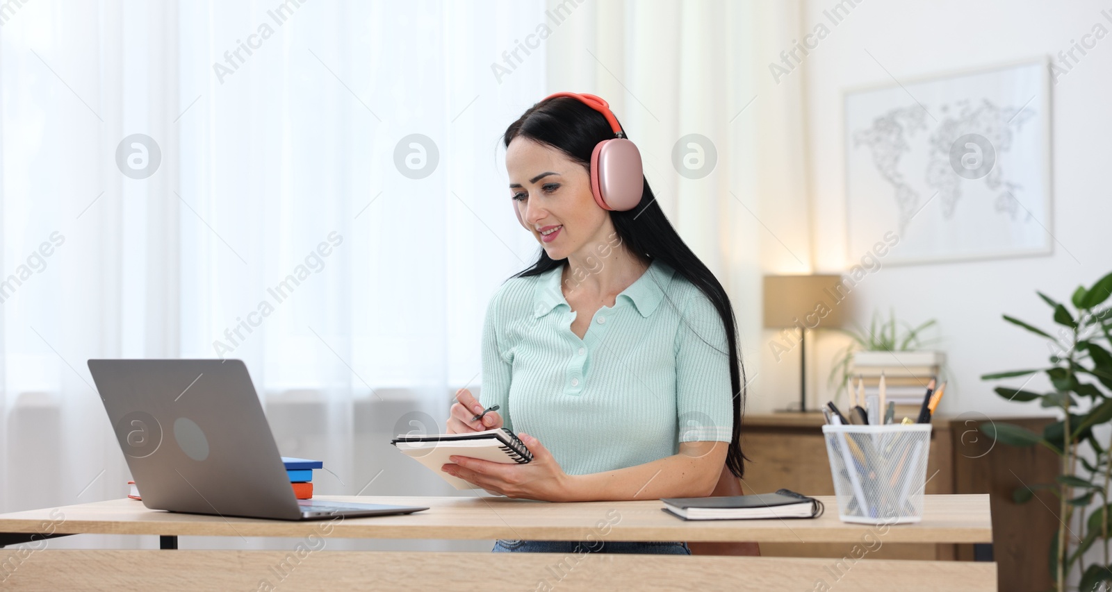 Photo of Woman in headphones taking notes while passing online test via laptop at table indoors