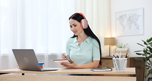 Photo of Woman in headphones taking notes while passing online test via laptop at table indoors
