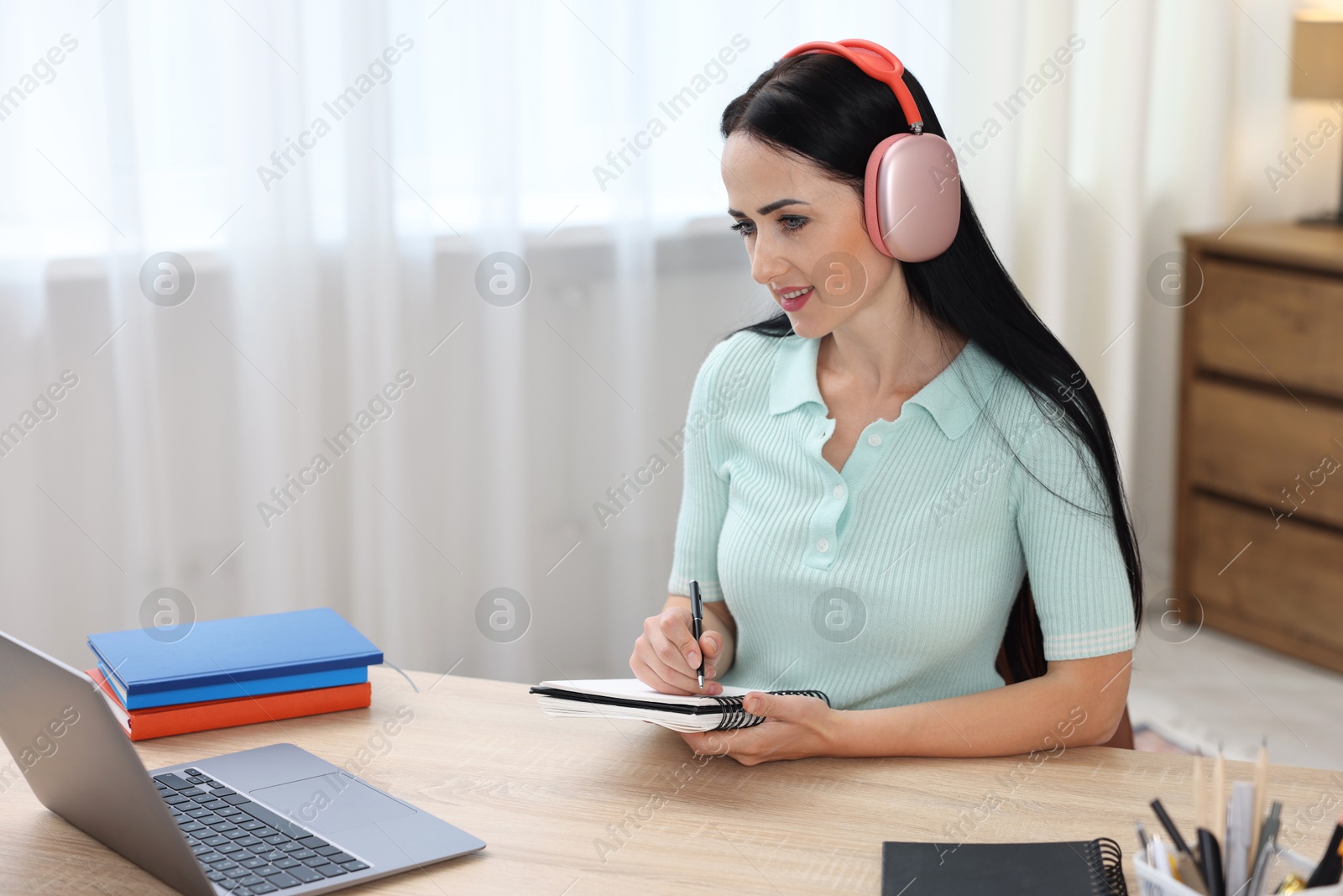Photo of Woman in headphones taking notes while passing online test via laptop at table indoors