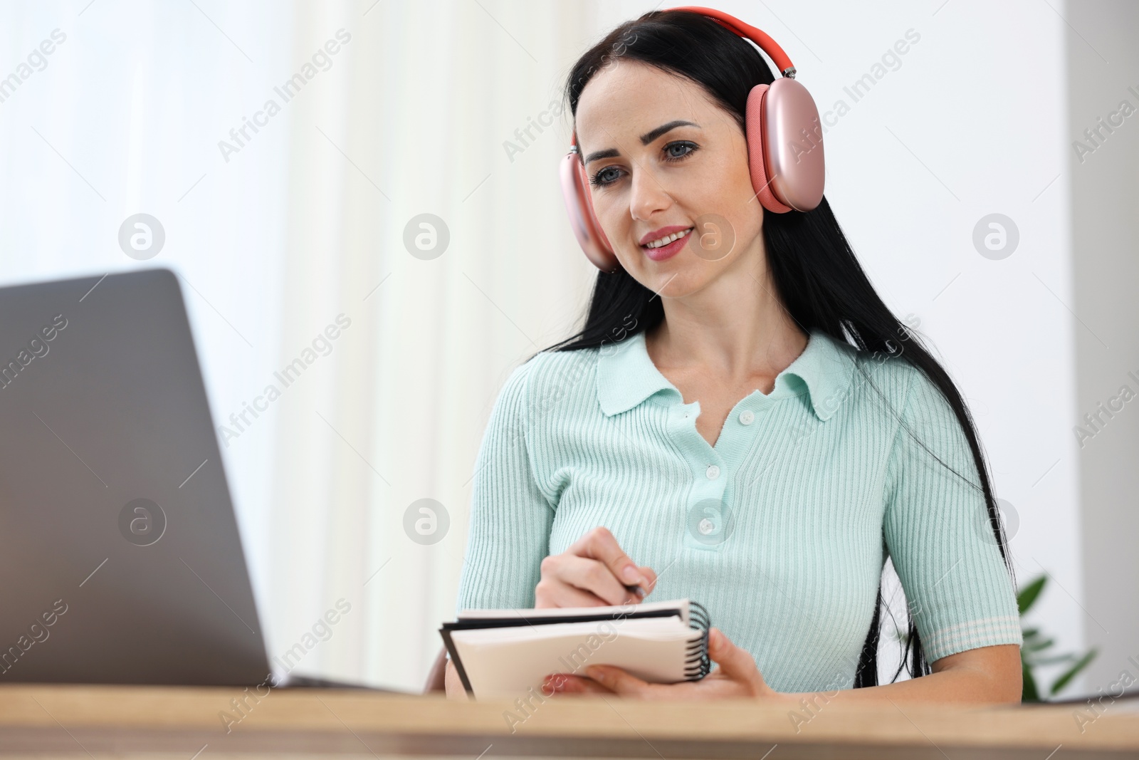 Photo of Woman in headphones taking notes while passing online test via laptop indoors