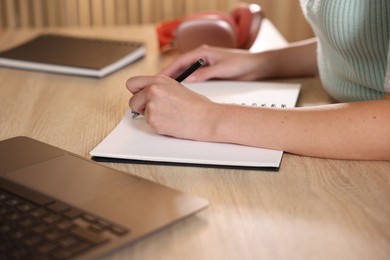 Photo of Woman taking notes while passing online test via laptop at wooden table indoors, closeup