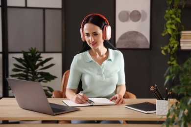 Photo of Woman in headphones taking notes while passing online test via laptop at table indoors