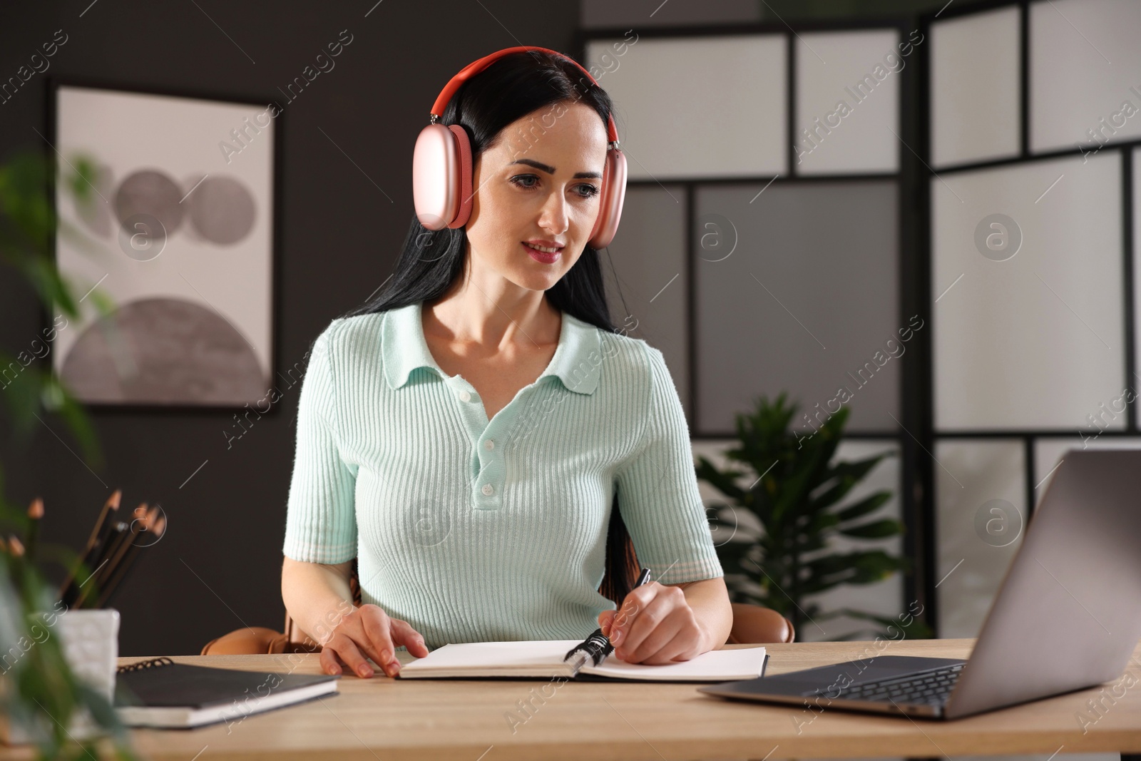 Photo of Woman in headphones taking notes while passing online test via laptop at table indoors