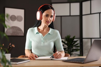 Photo of Woman in headphones taking notes while passing online test via laptop at table indoors