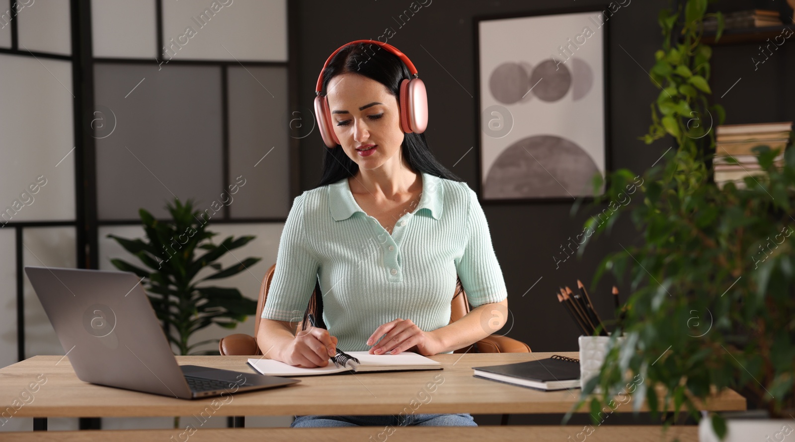 Photo of Woman in headphones taking notes while passing online test via laptop at table indoors