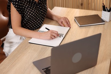 Photo of Woman taking notes while passing online test via laptop at table indoors, closeup