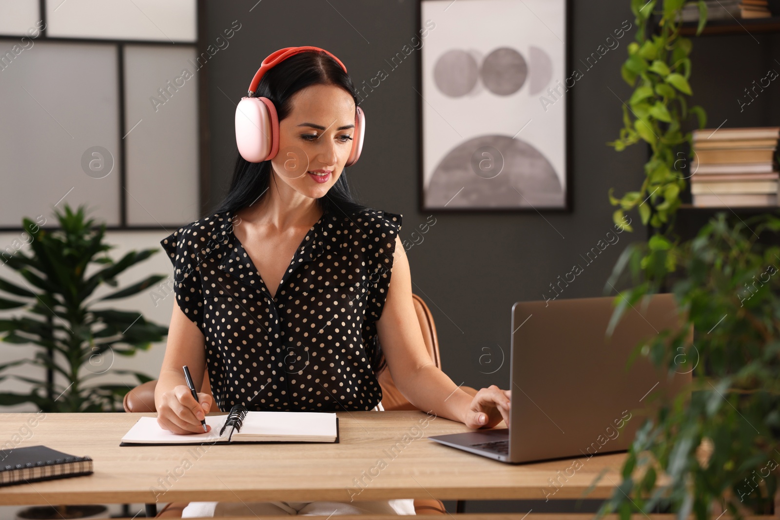 Woman in headphones taking notes while passing online test on laptop at table indoors Photo of Woman in headphones taking notes while passing online test on laptop at table indoors