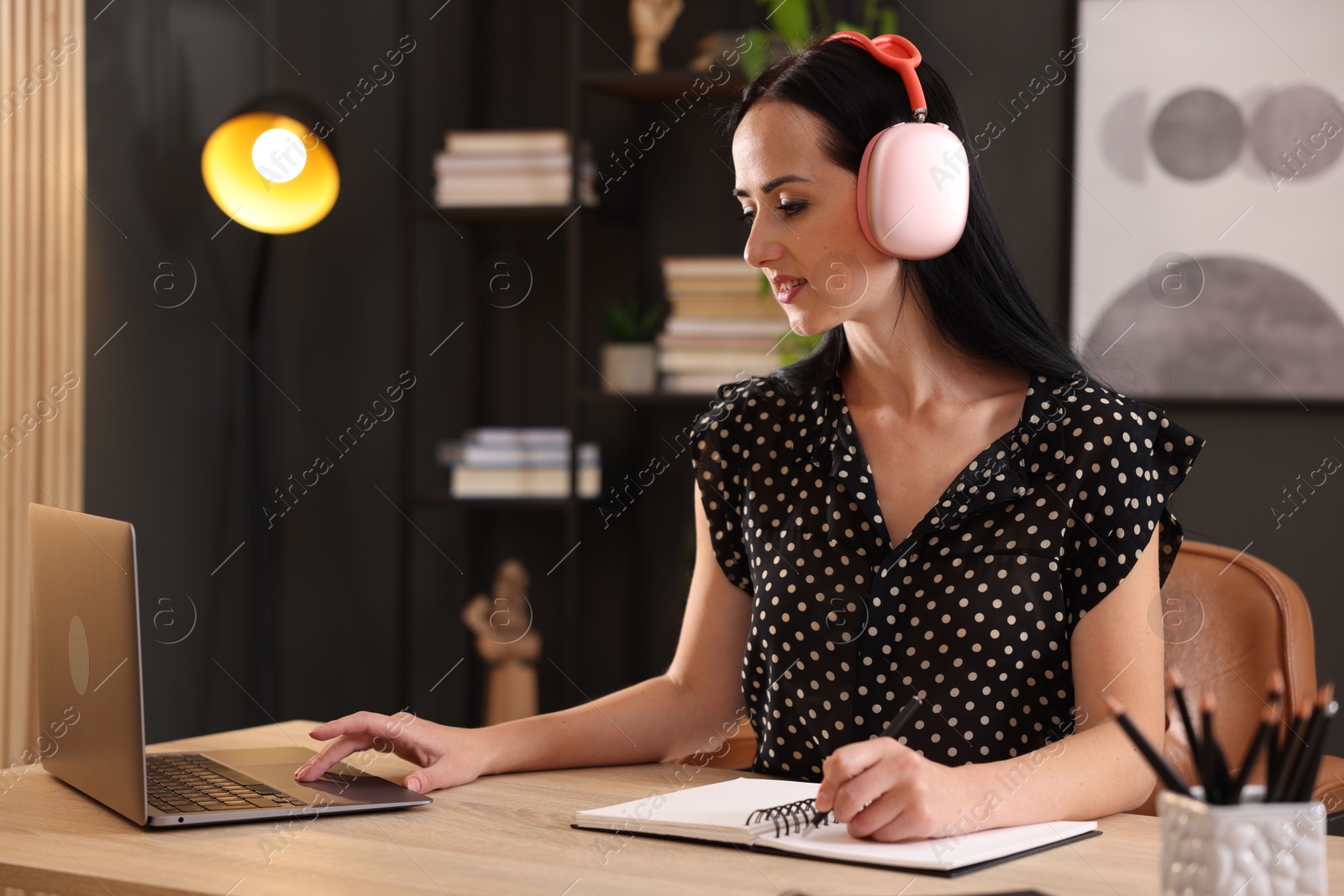 Woman in headphones taking notes while passing online test on laptop at table indoors Photo of Woman in headphones taking notes while passing online test on laptop at table indoors