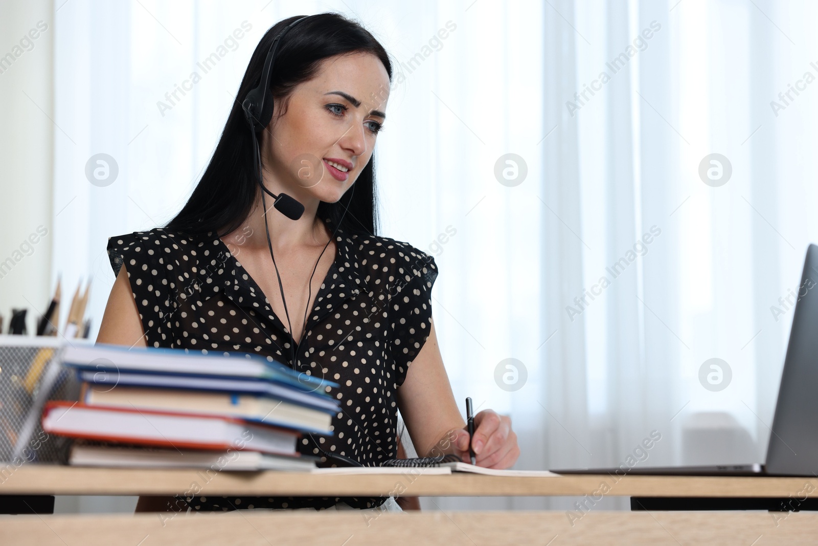 Woman in headset taking notes while passing online test via laptop at table indoors Photo of Woman in headset taking notes while passing online test via laptop at table indoors