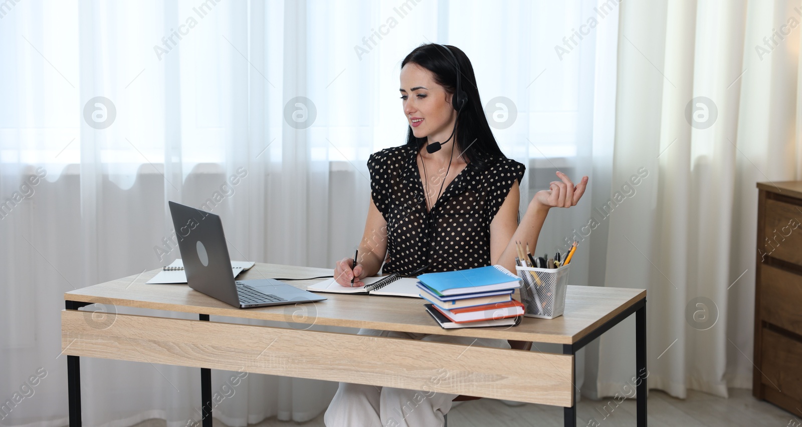 Woman in headset taking notes while passing online test via laptop at table indoors Photo of Woman in headset taking notes while passing online test via laptop at table indoors