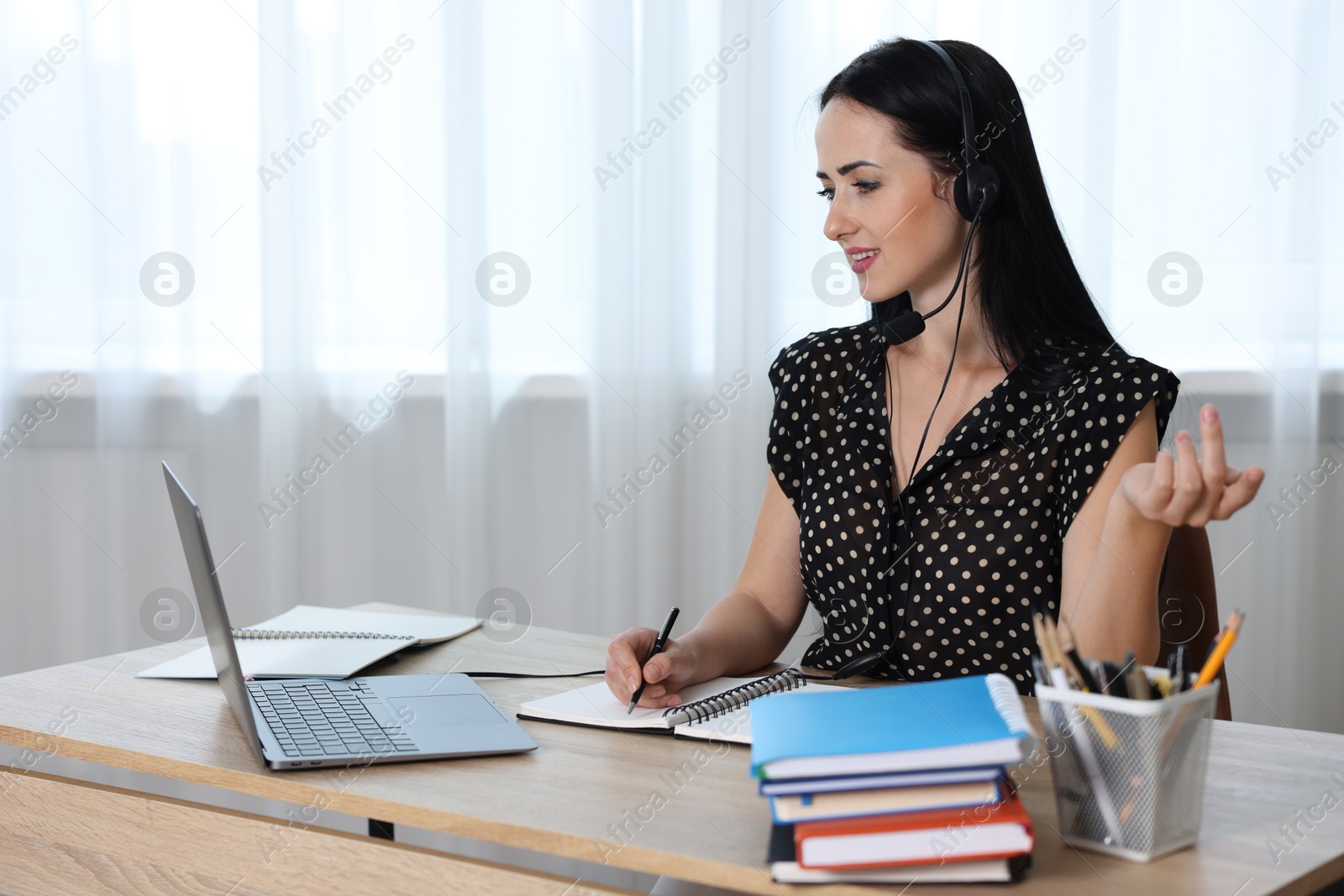 Woman in headset taking notes while passing online test via laptop at table indoors Photo of Woman in headset taking notes while passing online test via laptop at table indoors