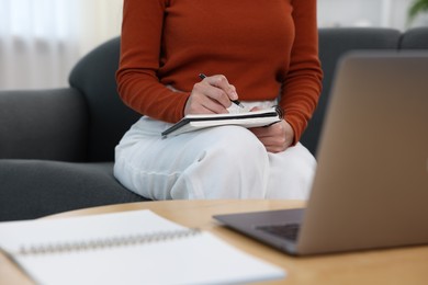Photo of Woman taking notes while passing online test via laptop at home, closeup
