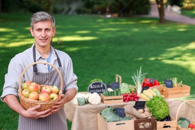 Photo of Happy farmer with apples in wicker basket near stall at market