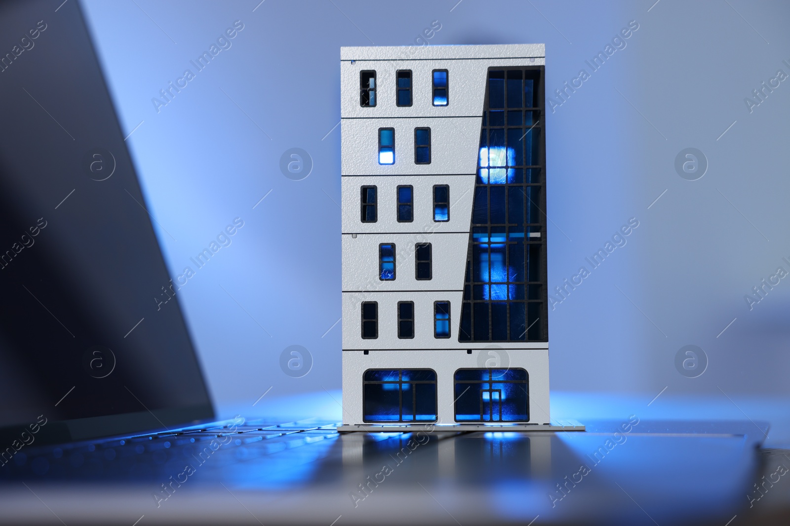 Photo of Real estate. Apartment building model and laptop on table in color light, closeup