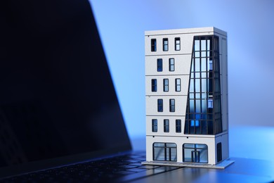Photo of Real estate. Apartment building model and laptop on table in color light, closeup