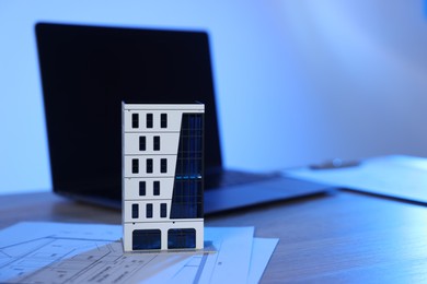 Photo of Real estate. Apartment building model, architectural drawings and laptop on table in color light, closeup