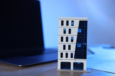 Photo of Real estate. Apartment building model and laptop on wooden table in color light, closeup