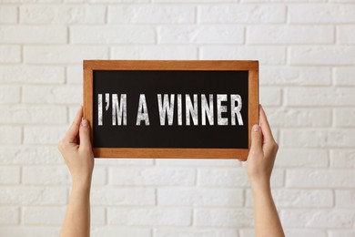 Image of Woman holding chalkboard with phrase I'm A Winner against white brick wall, closeup