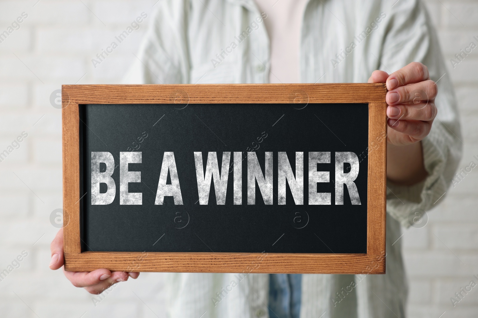 Image of Woman holding chalkboard with phrase Be A Winner against white brick wall, closeup