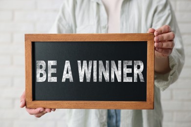 Image of Woman holding chalkboard with phrase Be A Winner against white brick wall, closeup