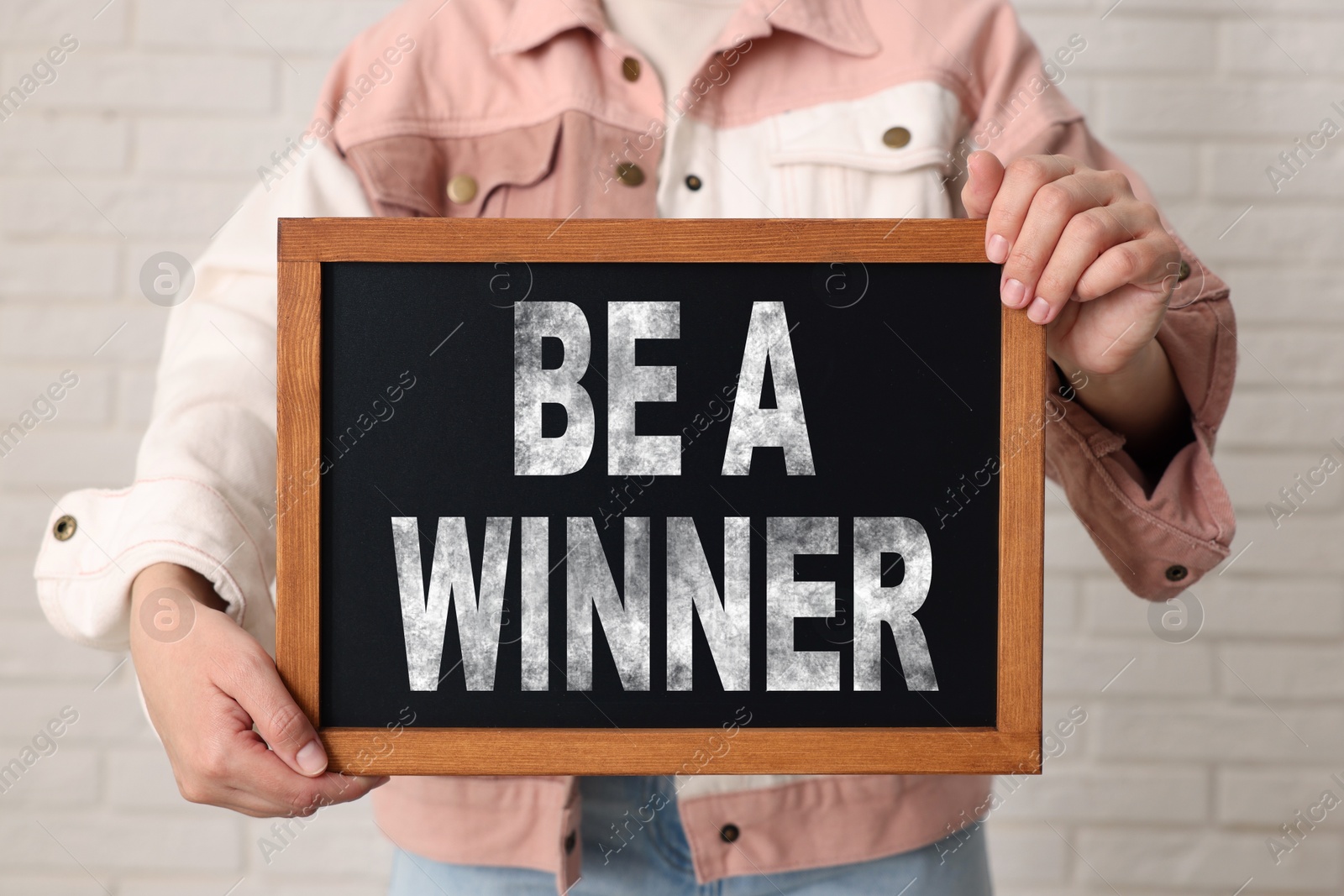 Image of Woman holding chalkboard with phrase Be A Winner against white brick wall, closeup