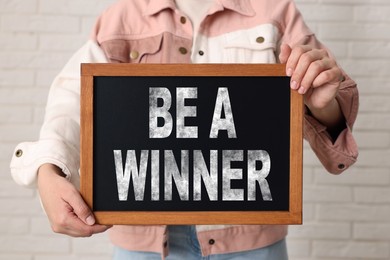 Image of Woman holding chalkboard with phrase Be A Winner against white brick wall, closeup