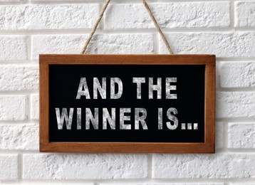 Image of Chalkboard with phrase And The Winner Is hanging on white brick wall