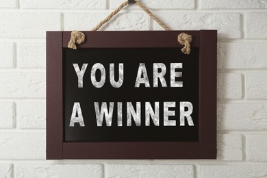 Image of Chalkboard with phrase You Are A Winner hanging on white brick wall