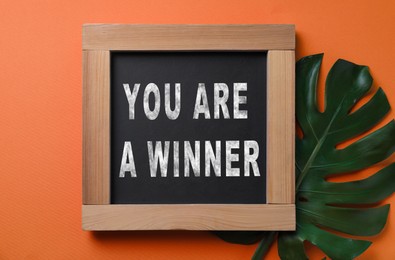 Image of Chalkboard with phrase You Are A Winner and green monstera leaf on dark orange background, top view