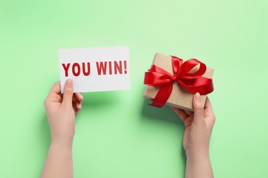 Image of Woman holding card with phrase You Win and gift box on light green background, top view