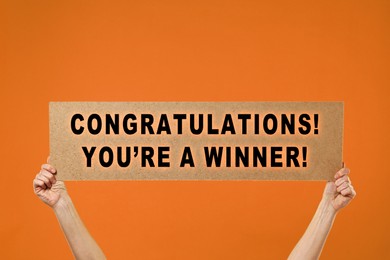 Image of Woman holding sign with phrase Congratulations! You're A Winner! against orange background, closeup