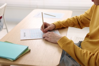 Photo of Student filling out answer sheet at wooden table indoors, closeup. Taking exam