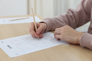 Photo of Student filling out answer sheet at wooden table indoors, closeup. Taking exam