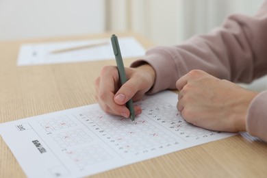 Photo of Student filling out answer sheet at wooden table indoors, closeup. Taking exam