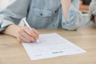 Photo of Student filling out answer sheet at wooden table indoors, closeup. Taking exam