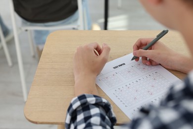 Photo of Student filling out answer sheet at wooden table indoors, closeup. Taking exam