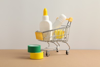 Photo of Glue in bottle, stick, small shopping cart and adhesive tapes on beige table against light grey background, closeup