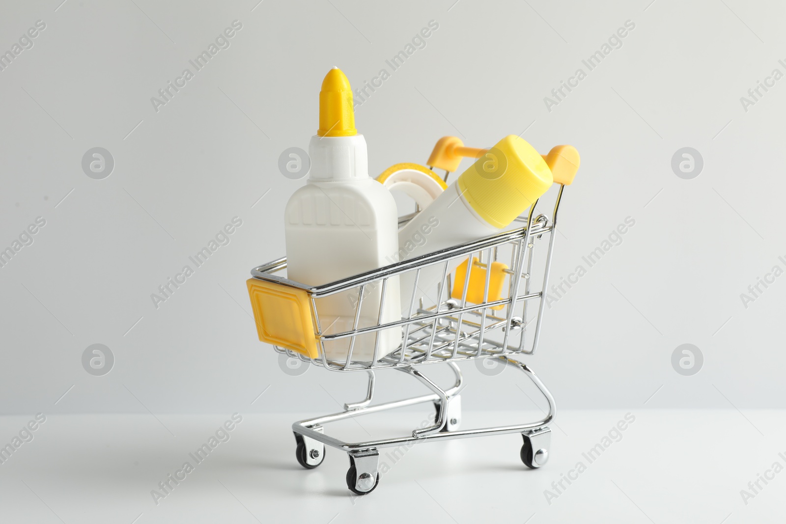 Photo of Glue in bottle, stick and adhesive tape in small shopping cart on light grey table, closeup