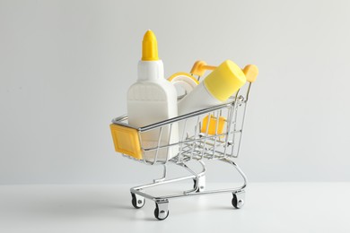Photo of Glue in bottle, stick and adhesive tape in small shopping cart on light grey table, closeup