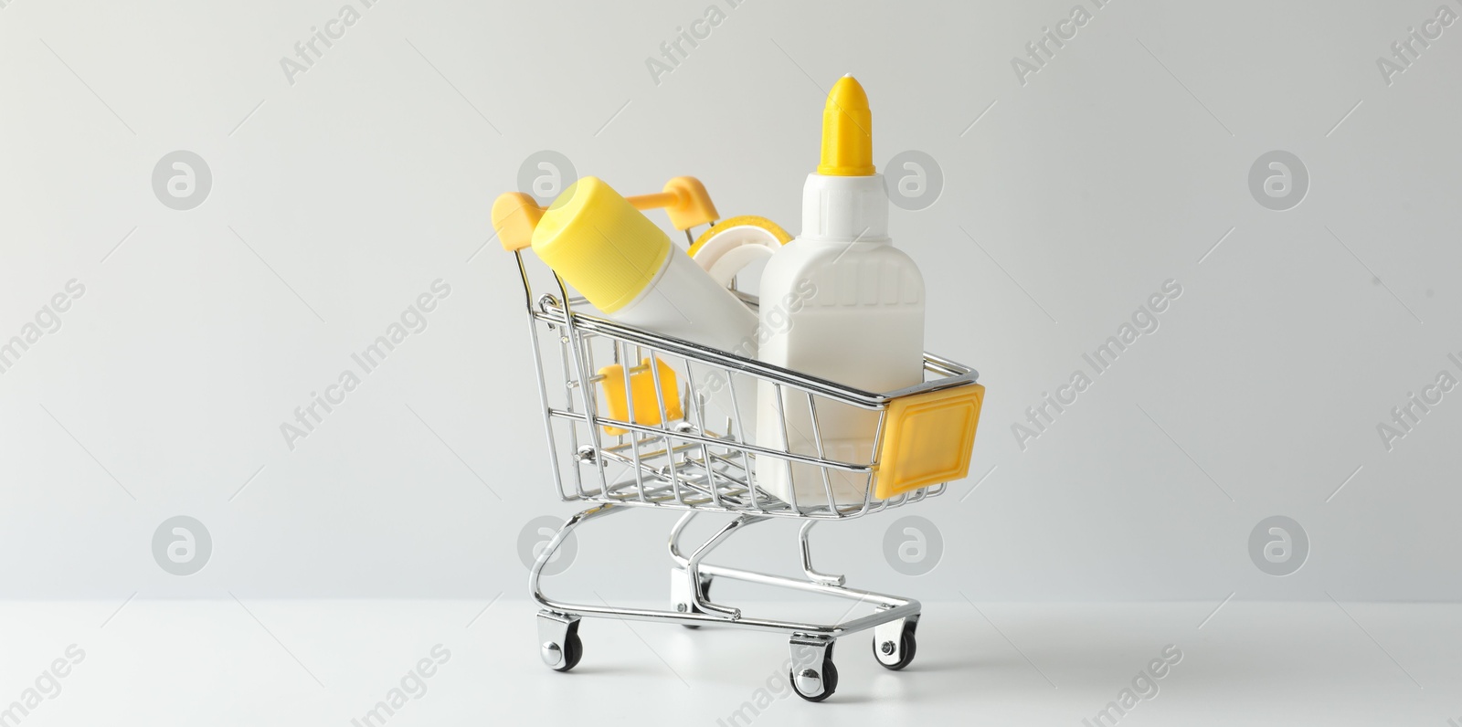 Photo of Glue in bottle, stick and adhesive tape in small shopping cart on light grey table, closeup