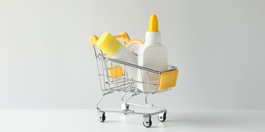Photo of Glue in bottle, stick and adhesive tape in small shopping cart on light grey table, closeup