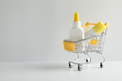 Photo of Glue in bottle, stick and adhesive tape in small shopping cart on light grey table, closeup. Space for text