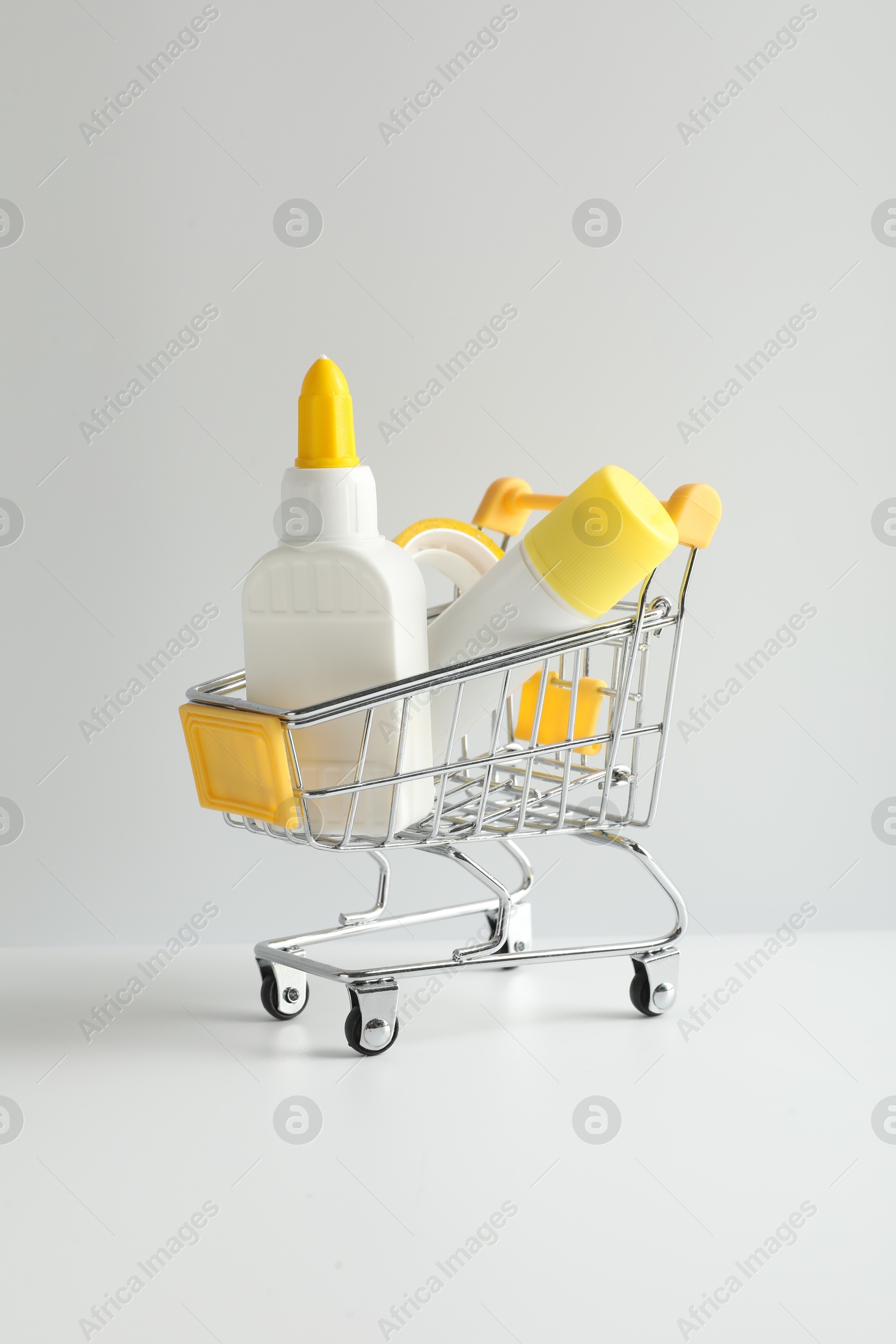 Photo of Glue in bottle, stick and adhesive tape in small shopping cart on light grey table