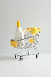 Photo of Glue in bottle, stick and adhesive tape in small shopping cart on light grey table