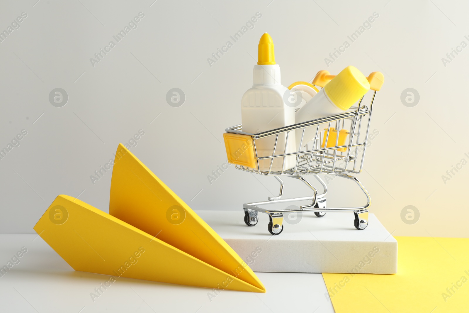 Photo of Glue in bottle, stick, adhesive tape in small shopping cart and yellow paper plane on light grey table, closeup