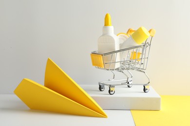 Photo of Glue in bottle, stick, adhesive tape in small shopping cart and yellow paper plane on light grey table, closeup
