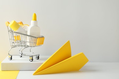 Photo of Glue in bottle, stick, adhesive tape in small shopping cart and yellow paper plane on light grey table, closeup. Space for text