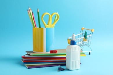 Photo of Glue in bottle, other stationery and small shopping cart on light blue background, closeup