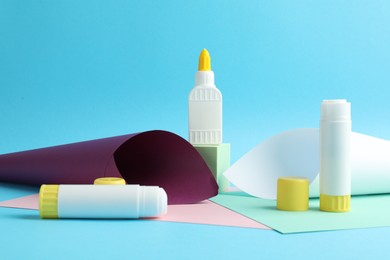 Photo of Glue in bottle, sticks and color paper on light blue background, closeup
