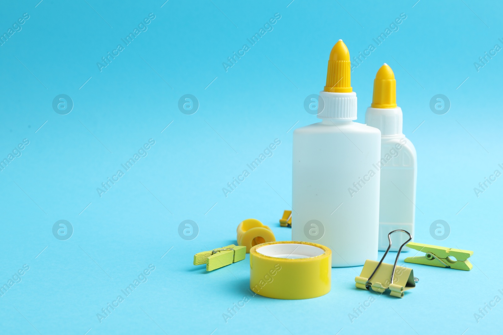 Photo of Glue in bottles, binder clip, sharpener and adhesive tape on light blue background, closeup. Space for text